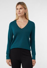 Kobieta z blond włosami, ubrana w turkusowy sweter w serek ze świecącymi wykończeniami i czarne spodnie, stojąca na tle jasnego, jednego koloru.