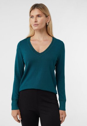 Kobieta z blond włosami, ubrana w turkusowy sweter w serek ze świecącymi wykończeniami i czarne spodnie, stojąca na tle jasnego, jednego koloru.