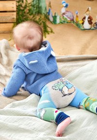 Gaiši zils adīts kapuce ar lāča ausīm, apvienots ar krāsainām legingām, kurās redzami dzīvnieku raksti. Fons ietver teksturētas segas.