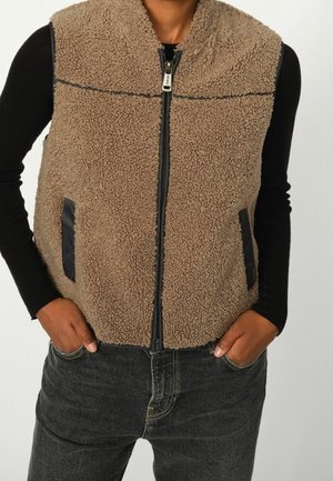 Personne portant une veste en polaire marron à fermeture éclair par-dessus un tee-shirt noir à manches longues, les mains dans les poches d'un jean gris foncé.