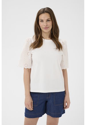 Donna con capelli castani lunghi indossa una camicetta bianca con maniche a sbuffo traforate a fiori e pantaloncini di denim blu scuro, in piedi contro uno sfondo bianco.