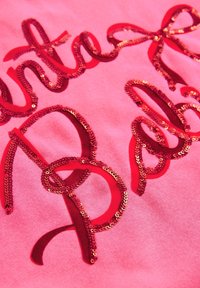 Rožinė medžiaga su raudonu, blizgučių puoštu tekstu, kuris sudaro stilizuotą raštą. Tekstūra pasižymi blizgančiais blizgučiais ir lygiu medvilniniu fonu.