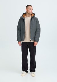 Mand iført grå polstret jakke over beige kabelstriktrøje, sorte bukser og hvide sneakers, står op ad en lys baggrund og ser mod højre.