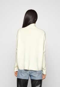 Jasna kremowa sweter z wysokim kołnierzem, plecioną teksturą i opuszczonymi ramionami, zestawiony z niebieskimi dżinsami i czarnymi skórzanymi spodniami.