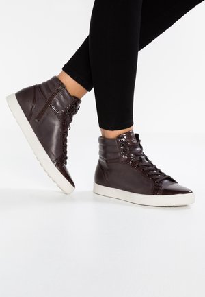 Person iført mørkebrune læder sneakers med højt skaft og hvide såler samt sorte leggings, stående på en hvid overflade.