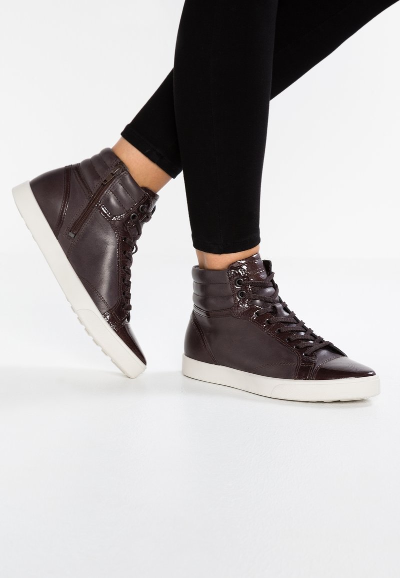 Personne portant des baskets montantes en cuir marron foncé avec des semelles blanches et des leggings noirs, debout sur une surface blanche.