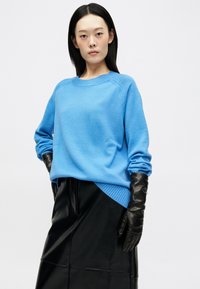 Ανοιχτό μπλε πλεκτό πουλόβερ με ραγκλάν μανίκια, στρογγυλή λαιμόκοψη και τελείωμα που καμπυλώνει προς τα πάνω από τη μία πλευρά. Συνδυάζεται με μια μαύρη δερμάτινη φούστα.