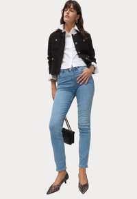 Chaqueta negra texturizada con botones dorados, camisa blanca, jeans de mezclilla azul claros y tacones de leopardo de punta, llevando un pequeño bolso negro.