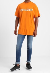 Camiseta naranja de manga corta con texto blanco, combinada con jeans de mezclilla en degradado y zapatillas deportivas negras. Conjunto casual y moderno.