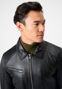 Junger Mann mit dunklem, welligem Haar, der eine schwarze Lederjacke über einem olivgrünen Rollkragenpullover trägt und seitlich vor einem schlichten Hintergrund schaut.