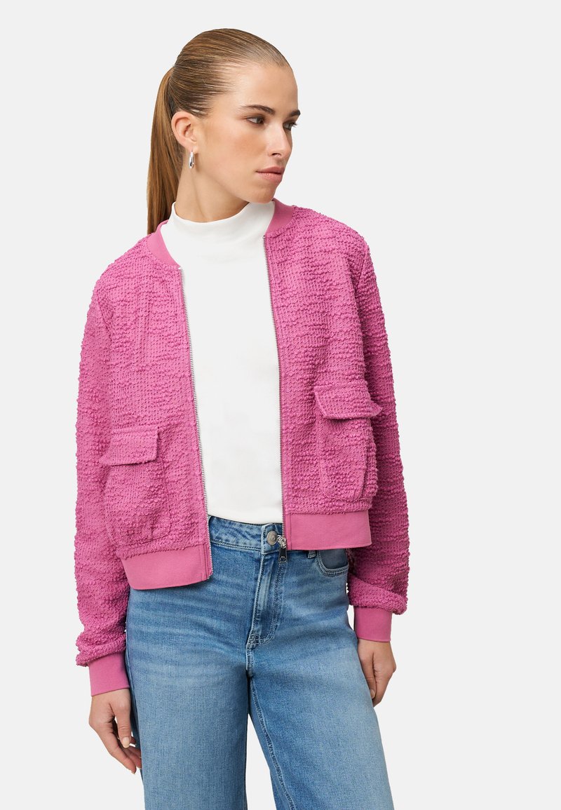 Veste bomber rose duveteuse avec poignets et ourlet côtelés, dotée de deux poches avant, portée sur un col roulé blanc et un jean bleu.
