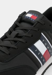 Zwarte sportieve schoen met een gestructureerd materiaal, vlakke veters en een logo-opdruk. Gestreept ontwerp met marineblauw, wit en rode accenten aan de zijkant.