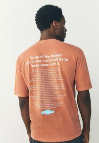 Bronzefarvet bomuld t-shirt med hvid tekst på ryggen, samt produktinformation og grafik nederst, der giver et vintage-look.