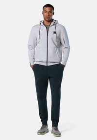Felpa leggera grigio chiaro con zip e cappuccio, due tasche frontali e dettagli verdi, abbinata a pantaloni joggers verde scuro e sneakers grigie.