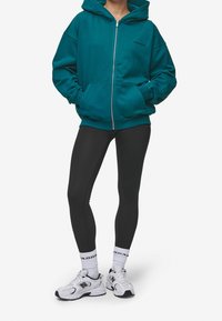 Felpa zip-up color teal con un grande cappuccio, tasche laterali, abbinata a leggings neri e sneaker bianche. La felpa presenta un logo sul petto.