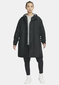 Cappotto impermeabile nero con cappuccio, maniche lunghe e chiusura a zip. Indossato sopra una maglietta grigia e pantaloni neri, completato da sneakers bianche.