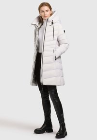 Padded white long coat with a hood, featuring a zip closure and black accents. Worn with black over-the-knee boots and fitted black pants.
