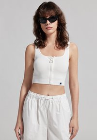 Crop top bianco a coste con zip frontale e scollatura quadrata. Abbinato a pantaloni corti bianchi larghi. Modella con occhiali da sole neri.