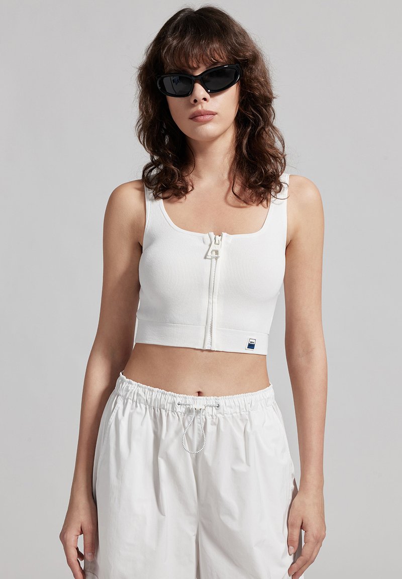 Crop top bianco a coste con zip frontale e scollatura quadrata. Abbinato a pantaloni corti bianchi larghi. Modella con occhiali da sole neri.