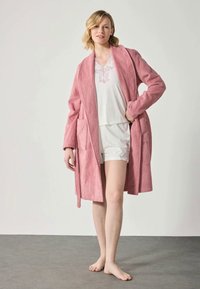 Rosa frottébadekåpe med teksturert stoff og to lommer, brukt over en hvit t-skjorte med korte ermer og shorts. Enkel og komfortabel design.