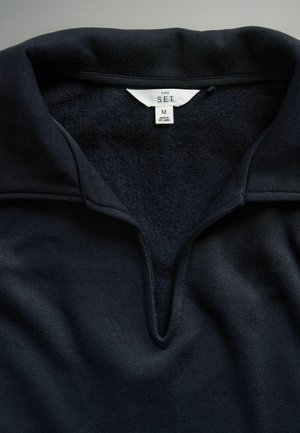 Sötét tengerészkék pulóver v-nyakú gallérral, puha tapintású, és méretjelző címkével, M méretű, Srí Lankán készült.