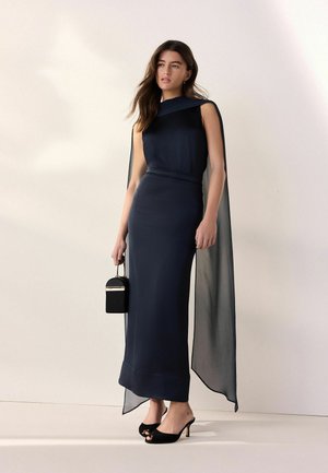 Nainen pukeutuneena hiukseton tummansiniseen iltapukuun, jossa on läpikuultava viitta, kädessään pieni musta käsilaukku ja jalassaan mustat korolliset mules-kengät.