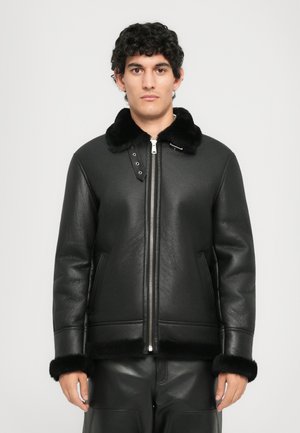 Man wearing black leather jacket with black fur collar, cuffs, and hem, standing against a plain light background.