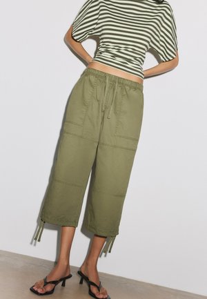 Donna che indossa pantaloni larghi corti verde oliva con orlo con coulisse, top a maniche corte a righe e sandali neri con tacco, in piedi contro un muro bianco.