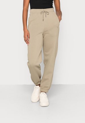 Persona con pantalones jogger beige con cordón y zapatillas blancas, de pie con una mano en el bolsillo contra un fondo liso.