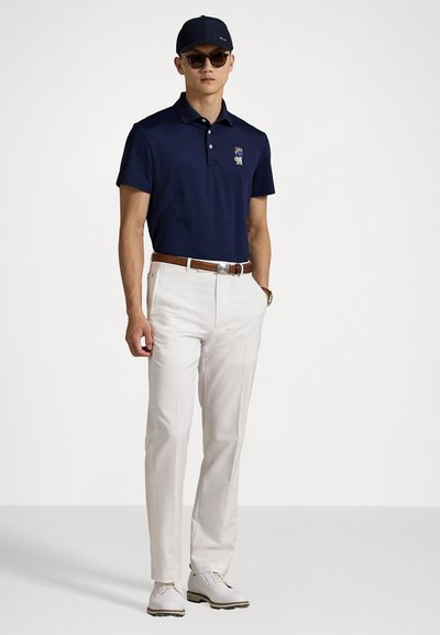 Tricou polo bleumarin cu un mic emblemă, asortat cu pantaloni albi de costum și curea din piele maro. Pantofi albi și o șapcă închisă completează aspectul.