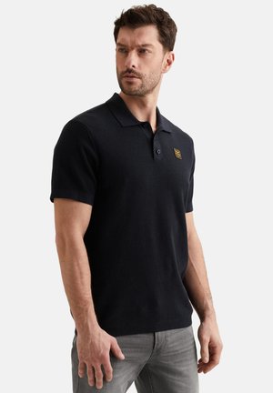 Mann trägt ein schwarzes Polohemd mit kurzen Ärmeln, Kragen und gelbem Logo-Patch, kombiniert mit grauer Hose, schaut nach links.