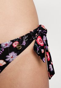 Parte de baixo de biquíni preta com estampa floral, com detalhe de laço amarrado, apresentando flores coloridas e um pequeno logótipo dourado. Textura de tecido suave.