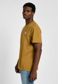 Kurzärmliges Senfgelb-T-Shirt mit klassischem Rundhalsausschnitt, versehen mit einem kleinen blau-weißen Etikett auf der Brust und einer glatten Baumwolltextur.