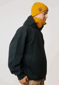 Czarna bluza z kapturem z kieszenią kangurką, zestawiona z prążkowaną musztardową czapką z naszywką z logo ze skóry. Jasne tło.