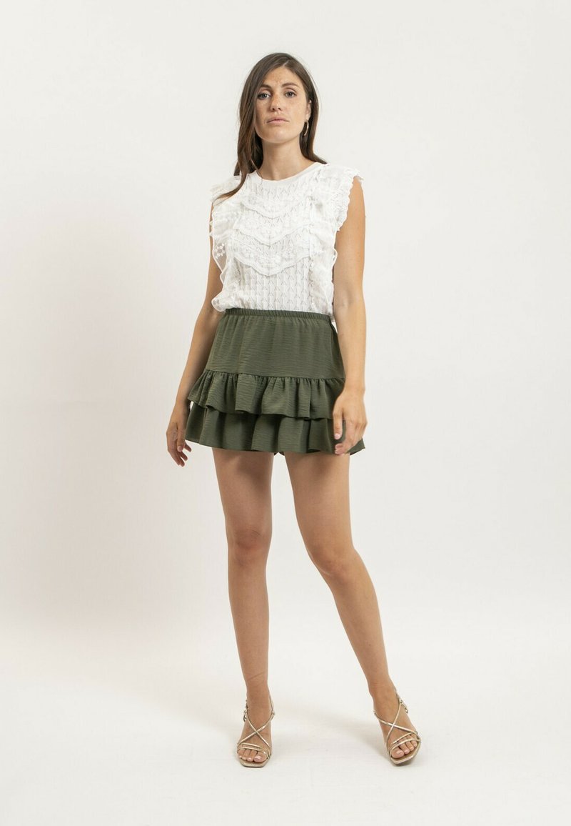 Blouse blanche à volants avec des détails texturés, associée à une jupe verte à volants. L'ensemble présente une taille ajustée et une silhouette évasée.