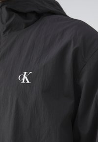 Giacca leggera nera con cappuccio, realizzata in tessuto testurizzato con un piccolo logo "cK" bianco sul petto. Design minimalista.