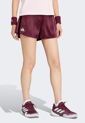Person iført bordeauxfarvede Adidas-shorts, matchende håndledsbind og bordeaux- og hvide Adidas-sneakers, stående mod en lysegrå baggrund.