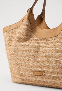 Sac fourre-tout en paille tressée de couleur beige naturel avec une surface texturée, doté d'une garniture en cuir tan et d'une petite étiquette de logo sur le devant.