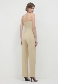 Beige trägerloser Jumpsuit mit tailliertem Oberteil, weitem Bein, seitlichen Taschen und glatter Textur. Kombiniert mit schwarzen High-Heels.