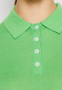 Zelené polo tričko s ribbovanou texturou, klasickým límcem a pěti odpovídajícími knoflíky vpředu. Vyrobeno z lehkého materiálu.