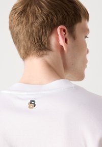 Valge puuvillane T-särk ümarate kraede ja väikese musta ja kuldse "B" logoga seljaosas, sile tekstuur ja puhas disain.