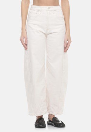 Donna che indossa jeans bianchi a gamba larga e vita alta e mocassini in pelle neri, mostrata dalla vita ai piedi su uno sfondo neutro.