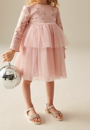 Jeune enfant vêtue d'une robe à manches longues rose à fleurs tenant une petite boule disco, portant des sandales scintillantes et debout sur un sol neutre.