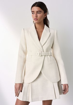 Femme portant un blazer texturé blanc cassé ceinturé avec des revers et une jupe plissée assortie, debout devant un fond clair uni.