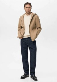 Beige lynlåsjakke over en cremefarvet sweater, kombineret med mørkeblå jeans og sorte sko. Jakken har en glat tekstur og et moderne snit.