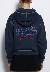 Granatowy sweatshirt z dużą czerwoną gwiazdą i haftowanym napisem "Bohaterowie". Posiada kontrastowe przeszycia oraz kaptur ze sznurkiem.