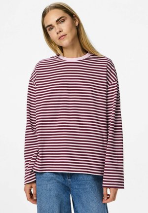 Mlada osoba s dugom plavom kosom, obučena u maroon i bijelu prugastu majicu dugih rukava i plave traperice, stoji uz bijelu pozadinu.