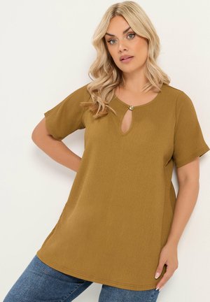 Blonde Frau trägt ein senfgelbes Kurzarmshirt mit Schlitz am Ausschnitt und blaue Jeans, steht vor weißem Hintergrund.