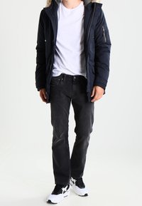 Parka aislante de color navy con capucha forrada de piel, bolsillos con cremallera, usada sobre una camiseta blanca y jeans negros con zapatillas blancas.