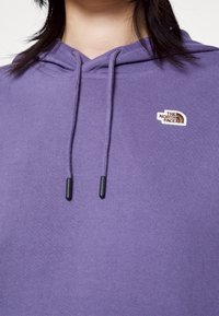 Gros plan d'une personne portant un sweat à capuche violet avec des cordons noirs et un petit logo The North Face blanc et marron sur la poitrine.